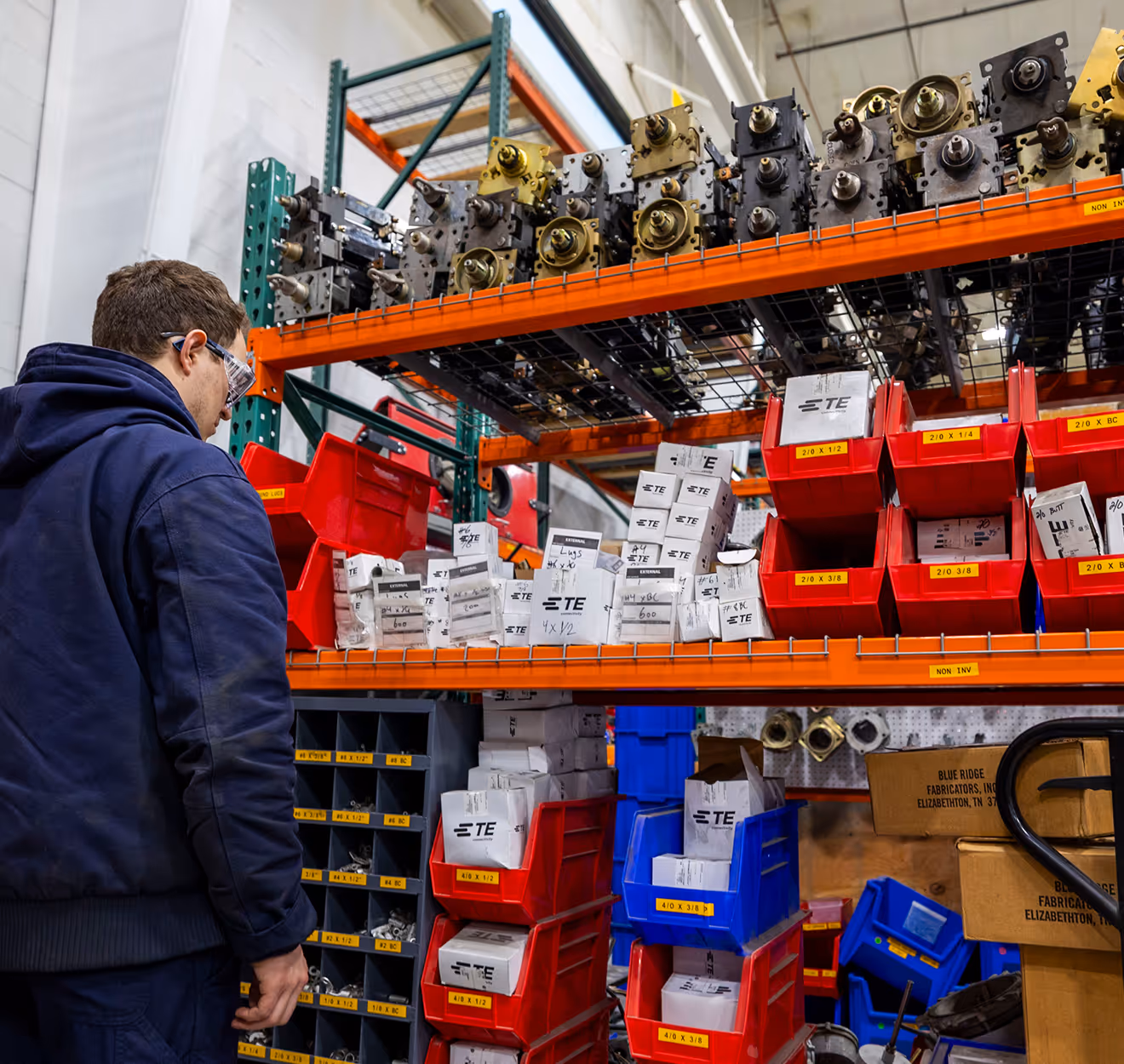 Man in safety glasses and blue jacket organizing labeled parts in red and blue bins on warehouse shelves.