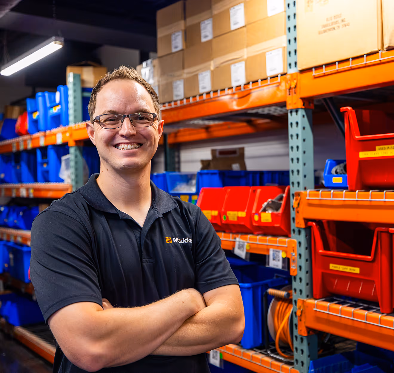 Smiling man with glasses in black Maddox shirt standing with folded arms in warehouse with red and blue bins.